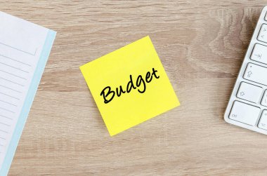 Bütçe metni sarı yapışkan bir nota üzerine yazılmış, beyaz bir bilgisayar klavyesi ve bir not defterinin yanındaki açık ahşap ofis masasına yerleştirilmiş..