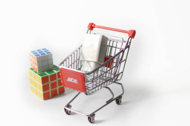 İki renkli Rubik küpünün yanında beyaz bir kutu taşıyan küçük bir alışveriş arabasının yaratıcı görüntüsü, eğlence alışverişini ve yaratıcılığı sembolize ediyor..