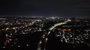 Bogor 'dan gelen şehir ışığı Jalan Tol Bogor üzerinde parlak bir zemin oluşturur, temiz şeritler, sabit trafik akışı ve yeşil ve gölgelerle çevrili huzurlu bir gece karayolu..
