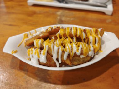 A close-up of crispy potato wedges generously topped with yellow cheese sauce and white mayonnaise or cream sauce