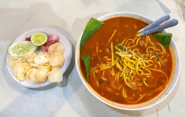 Baharatlı çorba ve sarı erişteyle dolu canlı bir kase Endonezya Mie Aceh yanında taze salatalık, limon, soğan turşusu ve emperyalist kraker..