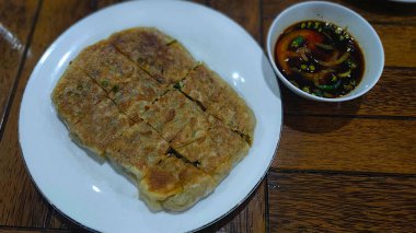 Taze, dilimlenmiş Martabak Kubang atau Martabak Mesir (lezzetli Endonezya gözlemesi) beyaz bir tabakta, soğanlı ve koyu ekşi soslu, ahşap bir masada servis edilir..
