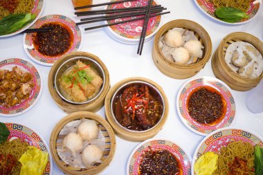 Çin Dim Sum ve şehriyelerinden oluşan bir ziyafet beyaz bir masaya yayılmış, bambu sepetleri, baharatlı soslar ve uzun çubuklar ile çeşitli buharda pişirilmiş hamur köfteleri sunuluyor..