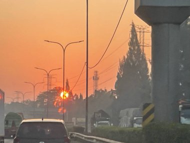 Canlı turuncu bir gün batımı arabaları, sokak lambaları ve elektrik hatları olan bir yol üzerinde parlıyor. Siluetli ağaçlar ve gökyüzüne karşı duran bir kule sisli ve sakin bir atmosfer yaratıyor..