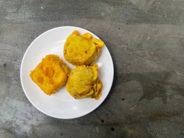 Altın hamurla kaplanmış üç çıtır tofu parçası beton bir masanın üzerinde beyaz bir tabakta servis ediliyor. Asya yemekleri, sokak atıştırmalıkları ya da mutfak temaları için mükemmel..