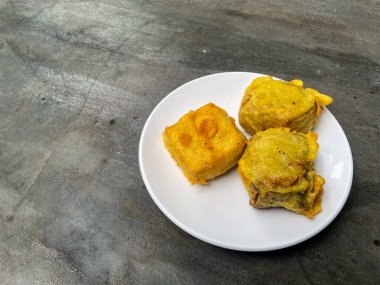 Hamur hamuruyla kaplanmış, çimento masasının üzerinde beyaz bir tabakta servis edilen üç altın çıtır tofu parçası. Asya yemekleri, sokak yemekleri ya da mutfak temaları için mükemmel..