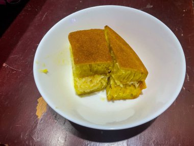 Popüler bir Endonezya tatlısı olan iki dilim tatlı peynir, beyaz bir tabakta oturur. Tabak kırsal ahşap bir masanın üzerinde, hafif soyulmuş bir yüzeyi var, rahat ve kırsal bir hissi vurguluyor..