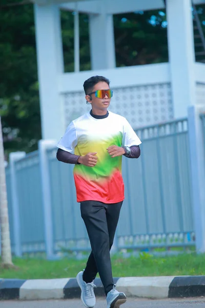 Aktif genç bir adam güneşli bir günde dışarıda koşuyor renkli spor kıyafetleri ve fütürist güneş gözlüğü takıyor.
