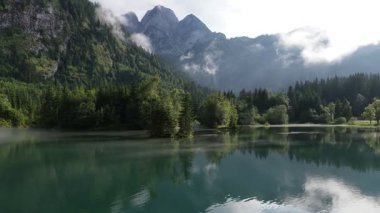 Sersemletici hava görüntüleri sisli bir sabahta sakin bir dağ gölünün üzerinde uçarken yemyeşil ormanın ve su yüzeyindeki alplerin görkemli zirvelerinin mükemmel yansımasını gösteriyor.