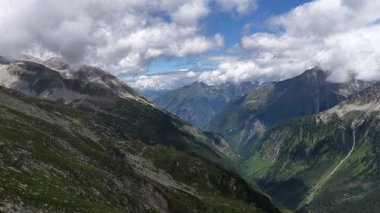 Görkemli hava perspektifi, yemyeşil yamaçları ve kayalık tepeleri olan, dinamik bulutlu mavi gökyüzü altında nefes kesici bir dağ vadisinde uçuyor. Saf doğal bir manzaranın ihtişamını yakalıyor.