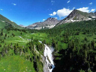 Beyaz su dalgalanan yeşil çimenler ve çamların arasından kayalık bir yamaçtan aşağı süzülür, berrak mavi gökyüzünün altındaki bir dağ vadisine süpürür, sakin ve el değmemiş bir çöl.