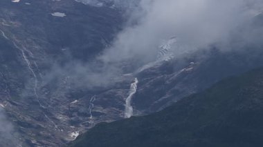 Kayalık bir dağda eriyen bir buzulun görkemli hava perspektifi, Alplerdeki sisli ve bulutlu bir arazide sarp yamaçtan aşağı akan çarpıcı bir şelale oluşturur.