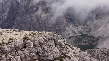 Dramatik hava görüntüleri, dolomitlerin kayalık yamaçlarında dinlenen büyük bir papağan sürüsünü gözler önüne seriyor. Görkemli, bulutlarla örtülmüş zirveler çarpıcı ve vahşi bir zemin oluşturuyor.