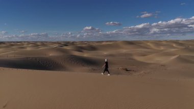 Mangystau, Kazakistan 'da engin ve kurak bir çöl arazisinde kumulun tepesinde yürüyen yalnız bir kadının hava görüntüsü dağınık bulutlarla dolu mavi bir gökyüzü altında keşif ve yalnızlığı sembolize ediyor.