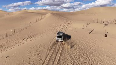 Yüksek açılı insansız hava aracı, altın kum tepecikleri üzerinde giden bir spor aracını izliyor. Kuru, kurak arazide, parçalı bulutlu bir gökyüzü altında lastik izleri gösteriyor. Maceracı bir çöl gezisi.