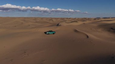 Mangystau, Kazakistan 'daki sarı kumullarda gezinirken engin bir çölde ilerleyen mavi bulutlu güzel bir gökyüzü altında uçan bir kamyonetin yüksek açılı görüntüsü.