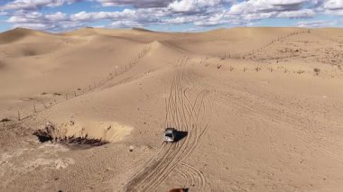 4x4 'lük bir arabanın yüksek açılı hava görüntüsü uçsuz bucaksız bir çöl arazisinde ilerlerken Hazar Denizi kıyısındaki altın kum tepelerinde, parçalı bulutlu bir gökyüzünün altında, macera ve keşifleri gösteren lastik izleri bırakıyor.