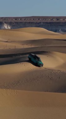 Yüksek açılı hava görüntüsü, mavi gökyüzü altında, Mangystau, Kazakistan gibi uzak beyaz kaya oluşumlarına sahip engin bir çölün altın ve dalgalı kum tepeciklerinde yol alan modern bir deniz aracını yakalıyor.