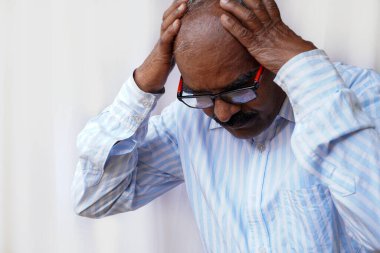 Yaşlı, gri saçlı, zarif tişörtlü yaşlı bir adam izole edilmiş beyaz arka planda dikiliyor. Baş ağrısı yüzünden çaresiz ve stresli. Çünkü acı ve migren. Ellerini başının üstüne koy..