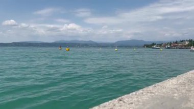 İtalya, Garda Gölü, 12 Temmuz 2025. Garda Gölü 'nün turkuaz suyu, palmiye ağaçları ve uzak dağlarla panoramik manzarası. Seyahat, doğa ve rahatlama videoları için mükemmel. Yazar: Djundiet A.