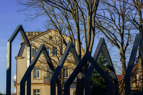 Çarpıcı siyah metal köşeli çerçeveler, Polonya 'nın huzurlu bir kasabasında, uzun çıplak ağaçları ve sıcak sonbahar ışığı olan klasik sarı tarihi bir binayı çerçeveleyerek, ön planda modern bir geometrik yapı oluşturur..