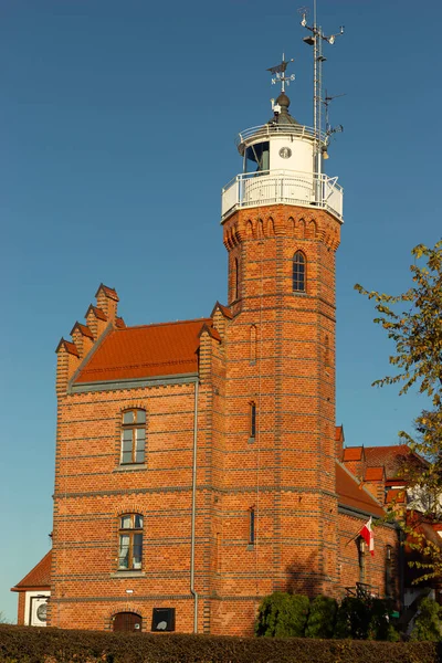 Polonya, Ustka 'daki büyüleyici kırmızı tuğlalı deniz feneri kulesi, tarihi binaya bağlanmış, açık mavi gökyüzü altında sıcak altın güneş ışığıyla yıkanmış, deniz cazibesiyle klasik sahil feneri.