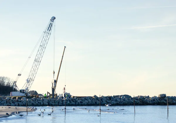 Ağır inşaat vinci ve kazıcılar Baltık Denizi kıyısında kayalık dalgakıranlar, kıyıda martılar, güvenlik yelekleri, endüstriyel liman gelişimi üzerinde çalışıyorlar.