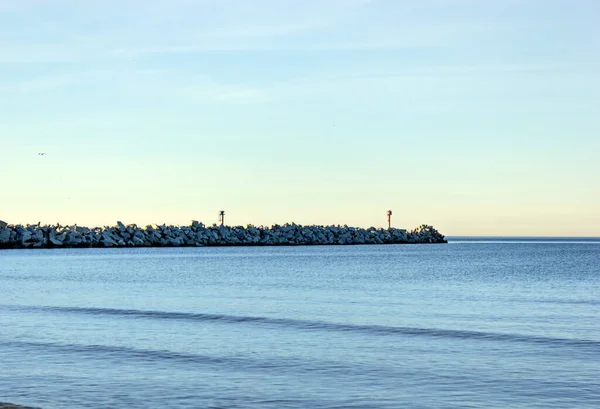 Sakin Baltık Denizi sularına uzanan uzun kayalık dalgalı deniz feneri yumuşak günbatımı gökyüzü altında, uzak kuşlar uçuyor, sakin kıyı ufku
