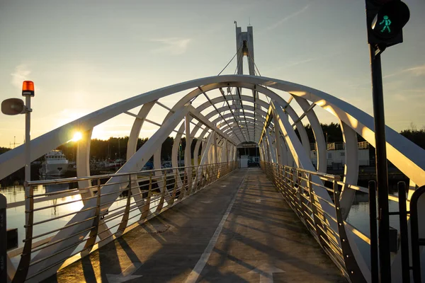 Altın gün batımında Ustka marinasındaki kemerli beyaz yaya köprüsüne bakıyorum. Parlayan yeşil sinyal, sakin liman suları, tekneler ve çam ormanları sahnesi sakin bir sahil manzarası yaratıyor.