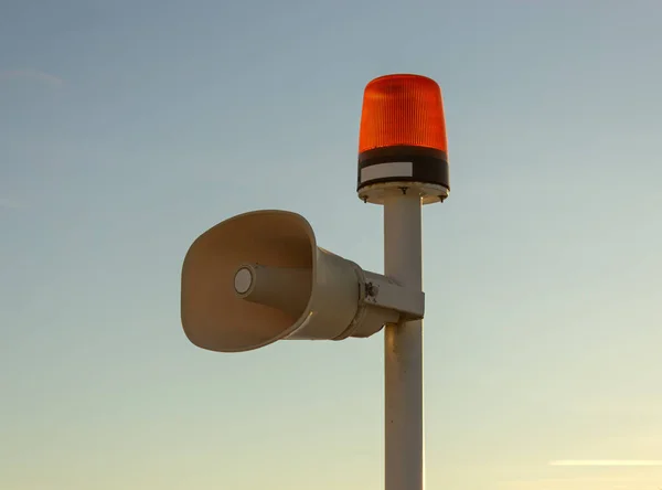 Ustka marinasında, deniz güvenliği ve liman atmosferini çağrıştıran sakin bir günbatımı gökyüzüne karşı beyaz bir kutba monte edilmiş turuncu dönen ışık ve bej hoparlörlü bir deniz uyarı fenerinin yakın çekimi.