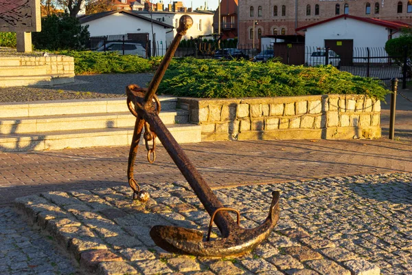 Günbatımında yeşillik ve eski tuğla binalarla çevrili Ustka şehir meydanındaki kaldırım taşı kaidesinde, tarihi, paslı çapa sergileniyor. Deniz mirası, turizm, deniz teması ve kıyı tarihi görselleri için ideal