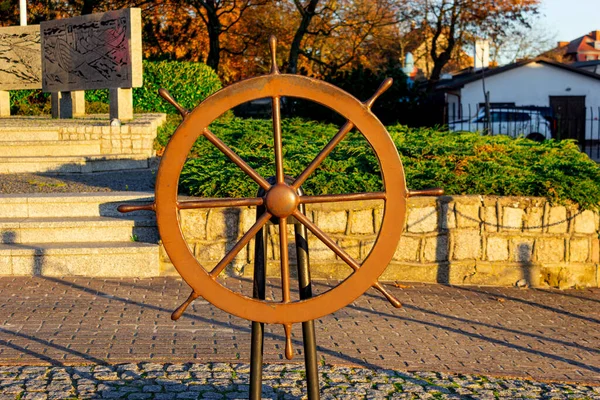 Ustka 'nın eski kasaba meydanındaki kaidede bulunan bronz geminin tekerlek heykeli, sıcak altın sonbahar ışığında yeşil ve tarihi binalarla çevrili. Denizcilik, deniz mirası, turizm ve denizcilik konuları için harika