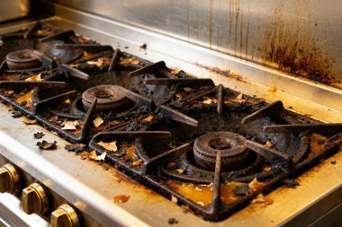 Bu resim çok kirli ve dağınık bir ticari mutfak sobasının yakın çekimini gösteriyor, temizlik ihtiyacını vurguluyor.