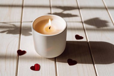 Yanan beyaz bir mum, küçük kırmızı kalp şekilleri ve yakındaki yaprakların gölgeleriyle çevrili, açık ahşap bir masada oturur.