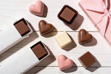 Kırsal bir ahşap yüzeyde sunulan kalp şekilleri ve kareler de dahil olmak üzere çeşitli çikolatalardan oluşan hoş bir koleksiyon büyüleyici pembe bir hediye kutusuyla birlikte.