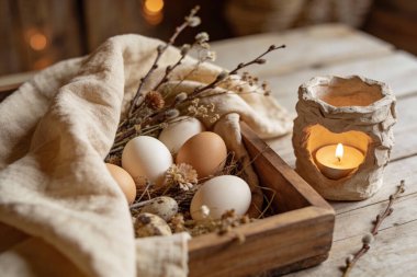 A rustic wooden crate filled with fresh eggs and pussy willow branches sits beside a warm, heart-shaped candle lantern, evoking a serene Easter atmosphere