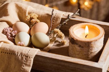 A warm and cozy Easter scene featuring natural colored eggs nestled in burlap beside a glowing candle and delicate dried floral accents
