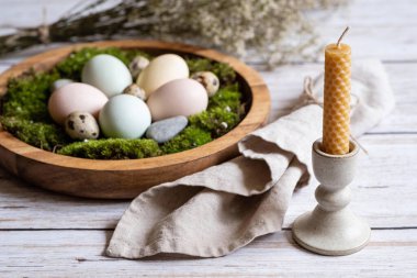 Canlı yeşil yosunların içine yerleştirilmiş pastel yumurtaların seramik bir tutucunun içindeki bal peteği balmumu mumuyla tamamlandığı kırsal bir Paskalya sofrası.