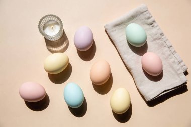 A collection of delicately colored Easter eggs are artfully scattered across a neutral surface, accompanied by a small lit candle and a folded linen napkin