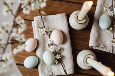 Zarif pastel yumurtalar, çiçek açan kiraz dalları ve kırsal bir ahşap masada mum yakan huzurlu bir Paskalya kutlaması sahnesi.