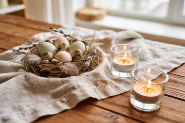 Bıldırcın ve tavuk yumurtalarıyla dolu bir kırsal Paskalya yuvası desenli keten kumaşın üzerinde iki yanan çay mumunun yanında oturur.