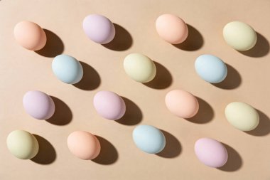 Pastel renkli Paskalya yumurtalarından oluşan bir kolleksiyon, bej rengi düz bir yüzeye ince gölgeler bırakarak simetrik bir desen içinde ustalıkla düzenlenmiştir.
