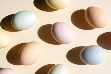Açık bej arkaplana saçılmış pastel renkli yumurtalardan oluşan bir derleme, yumuşak gölgeler bırakıyor.