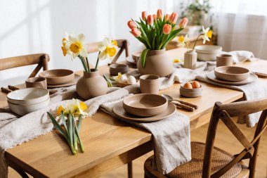 A beautifully arranged wooden dining table set for a meal with ceramic dishes, fresh flowers, and eggs, evoking a sense of springtime celebration and natural elegance