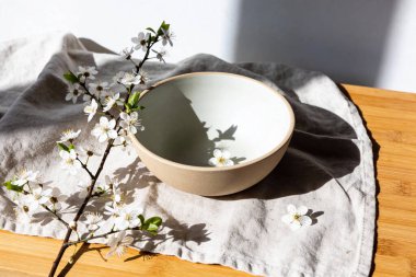 Basit bir seramik kase, beyaz kiraz çiçeklerinin açtığı bir dalla süslenmiş keten bir bezin üzerinde durmakta ve dingin bir bahar estetiği yaratmaktadır.