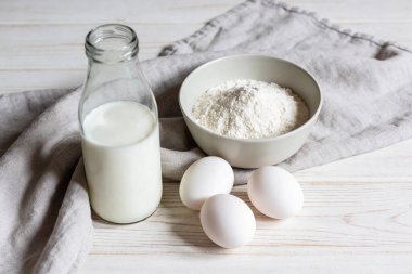 Bir şişe süt, bir kase un ve gri keten peçeteli açık ahşap bir yüzey üzerinde üç beyaz yumurta içeren temel pişirme malzemelerini içeren basit bir yaşam kompozisyonu.