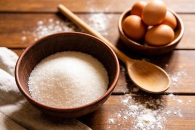 Kırsal bir kasede, bir tabak kahverengi yumurta ve ahşap kaşıkla birlikte, yakın plan bir şeker, hepsi un tozlu ahşap bir yüzey