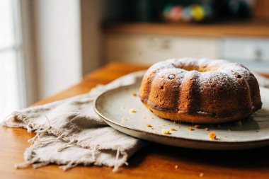 A freshly baked bundt cake, lightly dusted with powdered sugar, sits on a textured ceramic plate with crumbs scattered around