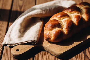 Güzel örülmüş bir challah somunu, katlanmış keten peçetenin yanında, kırsal ahşap bir yüzeyde, sıcak güneş ışığıyla yıkanır.