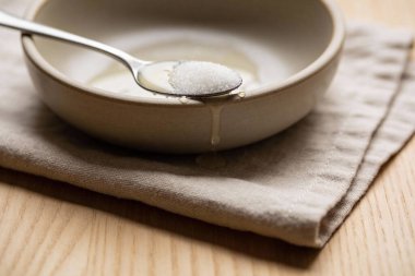 Metal kaşık beyaz şeker taneciklerini tutar, aşağıdaki seramik bir kaseye sıvı damlar, desenli keten bir bezin üzerinde dinlenir.